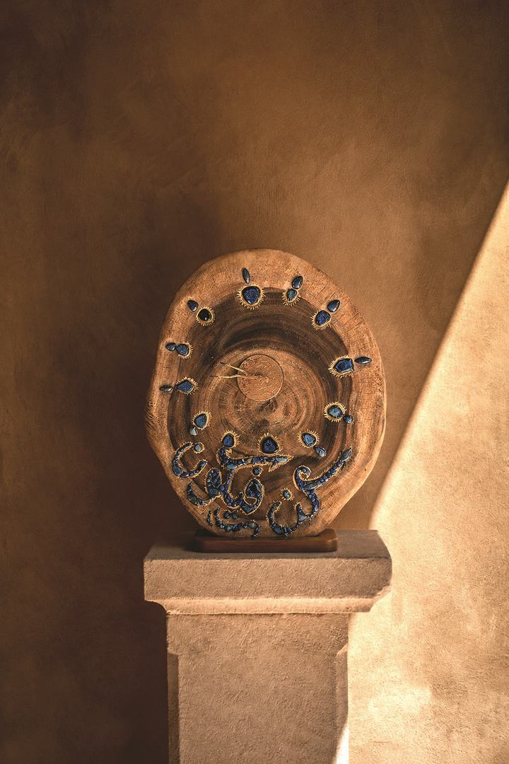 Round wooden piece with hand-carved Arabic calligraphy and inlaid blue stones, displayed on a stone pedestal in warm light