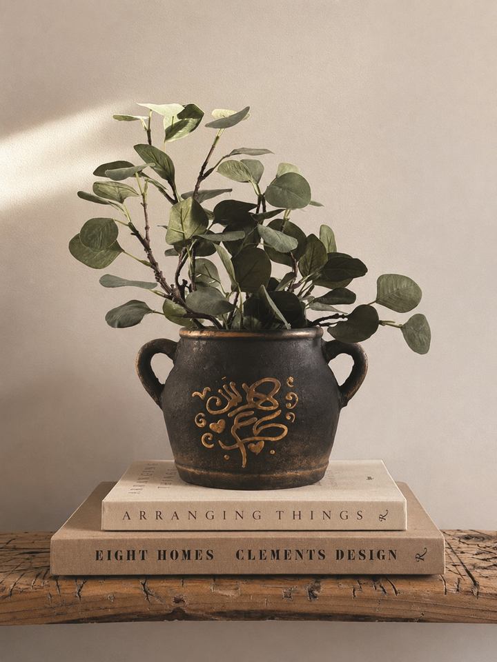 Dark pot with gold hand-lettered Arabic calligraphy holding a green plant, set on a stack of books