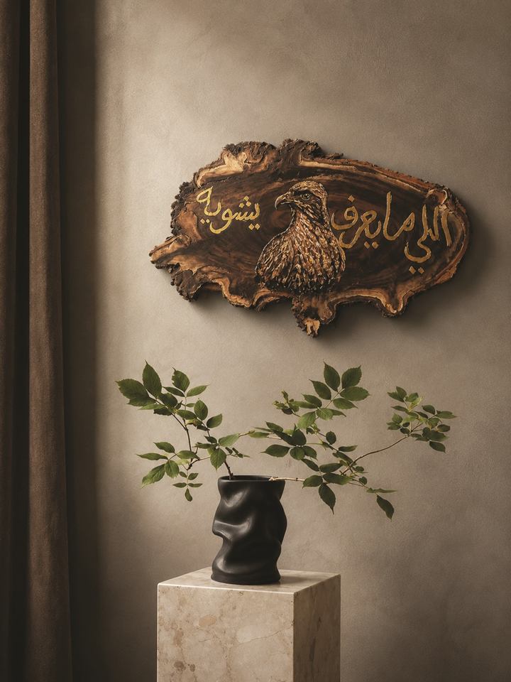 Live-edge wooden plaque with a carved falcon and hand-lettered Arabic phrase, hung on a textured wall
