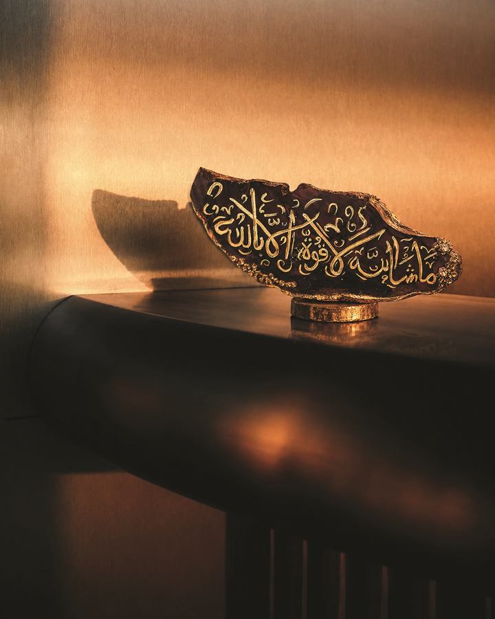 Engraved calligraphy piece reading ما شاء الله in gold on a dark carved surface, set on a small stand