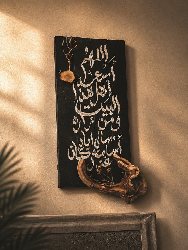 Black wooden panel hand-lettered with white Arabic calligraphy, finished with a piece of driftwood, on a sunlit wall